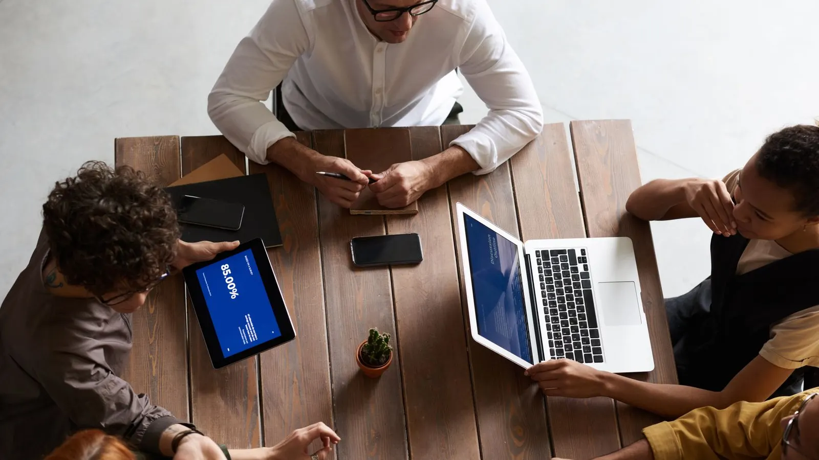 Overhead view of a team sharing tablets, laptops, and notes around a wooden table.