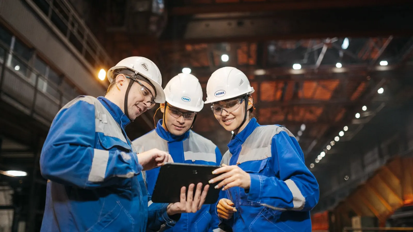 Three industrial colleagues in hard hats reviewing a tablet on a factory floor.