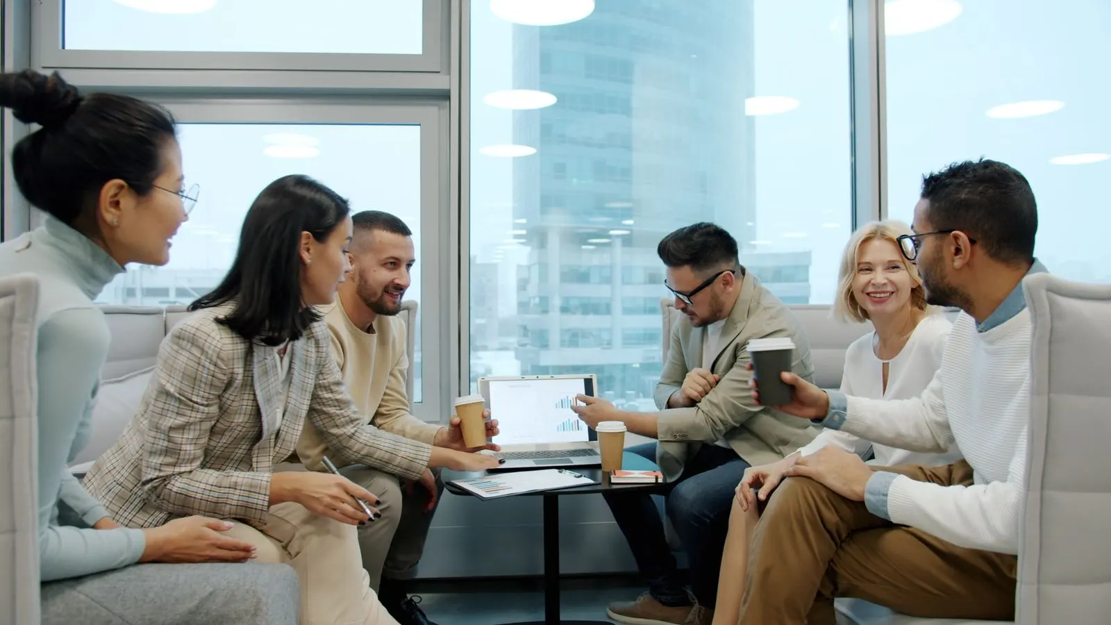 Six colleagues discussing work together around a low table in a bright office.