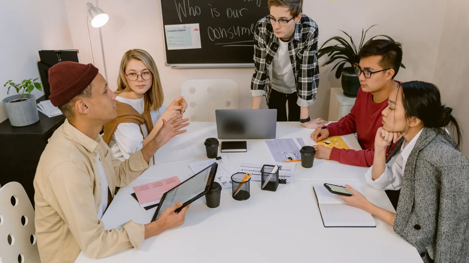 A standing colleague talking with a seated group around a white meeting table.