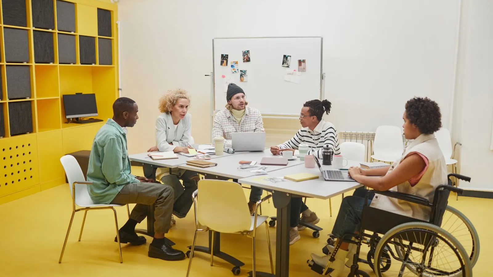 A group collaborating around a meeting table beside a yellow shelving wall.