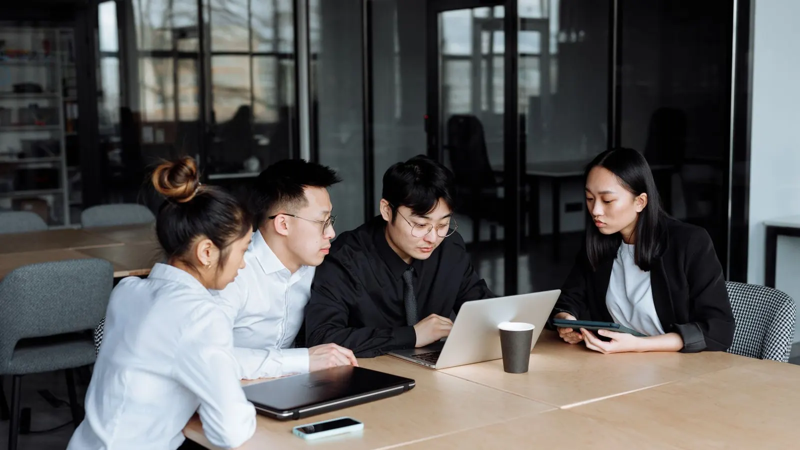 Colleagues gathered closely around a laptop in a glass-walled office.